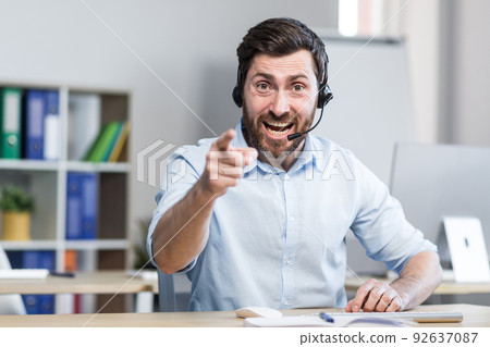 A young man, a consultant, an office worker, a manager talking on a video call in headphones, waving his arms, explaining something. Sitting at a computer desk in the office, in a white shirt 92637087