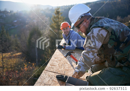Carpenter hammering nail into OSB panel on the roof top of future cottage. Man worker building wooden frame house. Carpentry and construction concept. Carpenter hammering nail into OSB panel on the roof top of future cottage. Man worker building wooden frame house. Carpentry and construction concept. 92637311