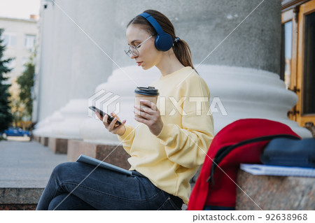 Portrait of casual student girl in headphones using mobile phone app outdoors. Young woman student 92638968