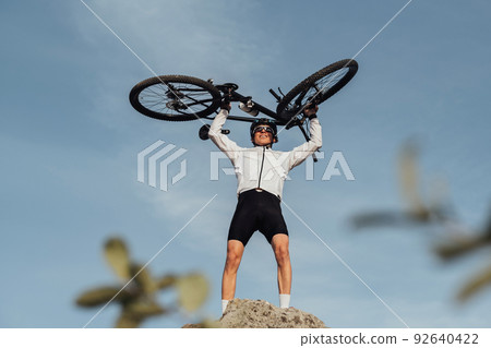 Professional Male Cyclist Raising Hands with Bicycle while Standing on the Top of Hill, Sky on the Background Professional Male Cyclist Raising Hands with Bicycle while Standing on the Top of Hill, Sky on the Background 92640422