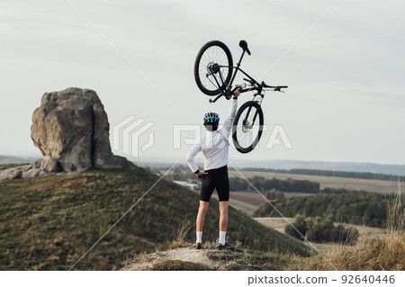 Professional Male Cyclist in Helmet Raising Hand with Bicycle while Standing on the Top of Hill, Giant Stone, Miracle of Nature on the Background 92640446