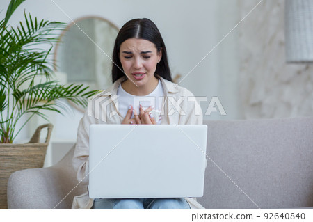 Young beautiful woman alone at home in depression crying, brunette sitting at home and using laptop for online consultation call with psychotherapist Young beautiful woman alone at home in depression crying, brunette sitting at home and using laptop for online consultation call with psychotherapist 92640840
