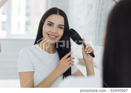 Beautiful young woman combing her hair in the morning, looking in a big round mirror and smiling, brunette in a bright bedroom at home Beautiful young woman combing her hair in the morning, looking in a big round mirror and smiling, brunette in a bright bedroom at home 92640967