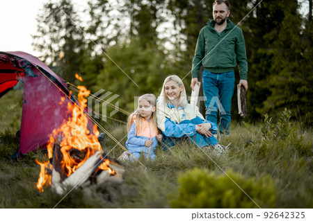 Young family sit by the campfire near tent on nature Young family sit by the campfire near tent on nature 92642325