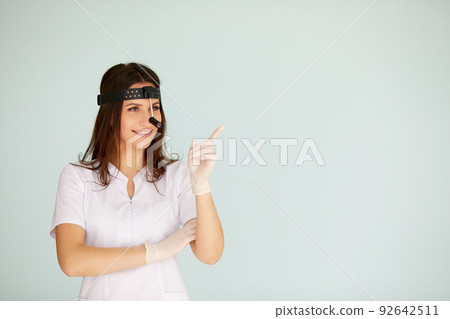 Cute young female otolaryngologist standing with illuminator for otolaryngologist on her head on bright background Cute young female otolaryngologist standing with illuminator for otolaryngologist on her head on bright background 92642511