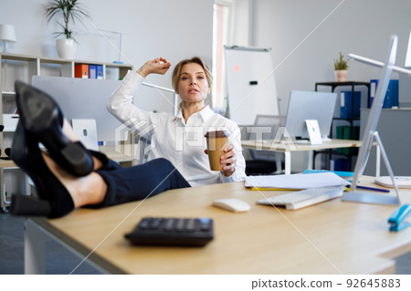 Rude female boss with a cup of coffee put her legs on the desk and throws crumpled paper at her worker 92645883