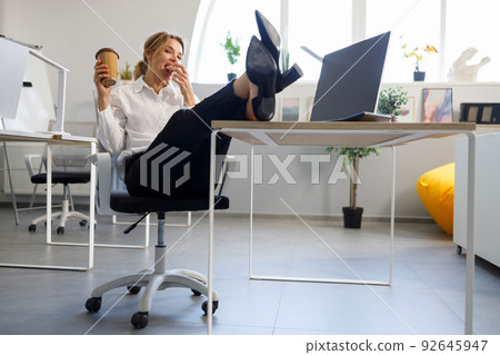 Business woman with a cup of coffee put her legs on the desk and eats cookies Business woman with a cup of coffee put her legs on the desk and eats cookies 92645947