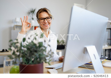 Middle-aged business woman sits at a computer in the office and shows ok gesture with her fingers Middle-aged business woman sits at a computer in the office and shows ok gesture with her fingers 92646413