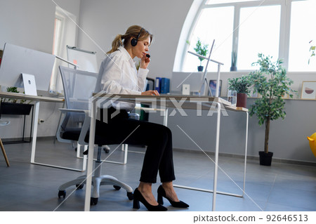 Female support center operator in headset talking with customer while sitting in office at computer 92646513