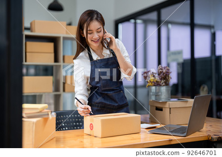 Business owner talking on mobile phone and taking order. Female asian entrepreneur working at home office confirming the order on phone. Business owner talking on mobile phone and taking order. Female asian entrepreneur working at home office confirming the order on phone. 92646701