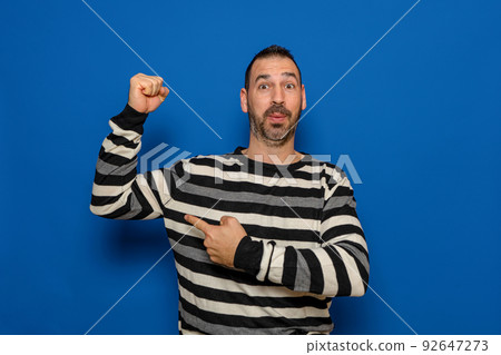 Unshaven 40 year old man in striped pullover pointing finger at biceps muscles demonstrating strength power isolated on pastel color background studio portrait. People lifestyle concept. 92647273