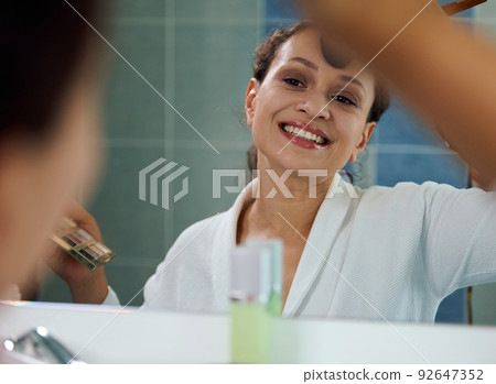Stunning woman applies mattifying powder on her face, satisfied of her appearance, looks at her reflection in the mirror 92647352