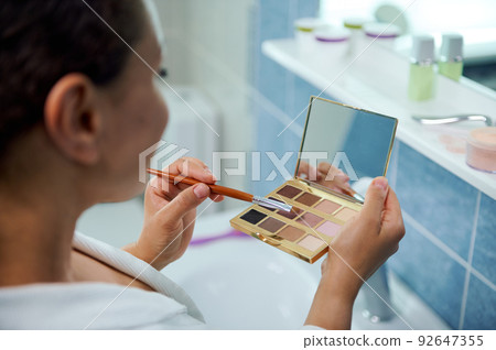 Rear view of a blurred woman holding eyeshadow palette with small cosmetic mirror and makeup brush, applying make-up 92647355