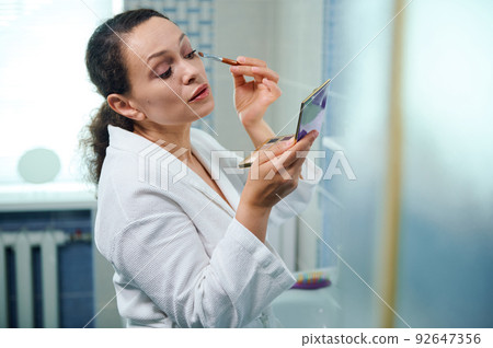 Beautiful Hispanic woman in a white bathrobe, holding a colorful eyeshadow palette and applying makeup on upper eyelids Beautiful Hispanic woman in a white bathrobe, holding a colorful eyeshadow palette and applying makeup on upper eyelids 92647356