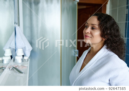 Self-confident 35s woman of natural beauty, in white bathrobe, enjoying fresh morning beauty procedures at home bathroom 92647385