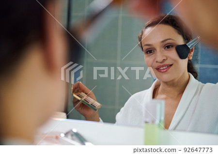 Delightful stunning young woman applies a mattifying powder on her face, satisfied of her appearance and natural make-up Delightful stunning young woman applies a mattifying powder on her face, satisfied of her appearance and natural make-up 92647773