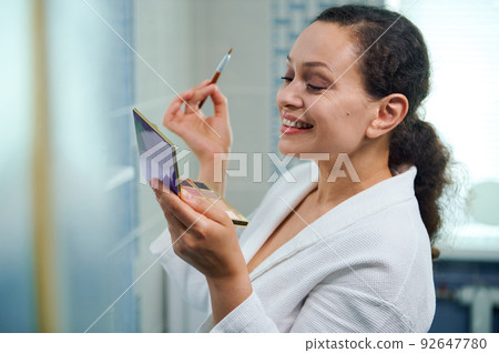 Stunning middle-aged woman, in bathrobe, holding a makeup palette, smiles while is applying eye shadows to upper eyelid 92647780