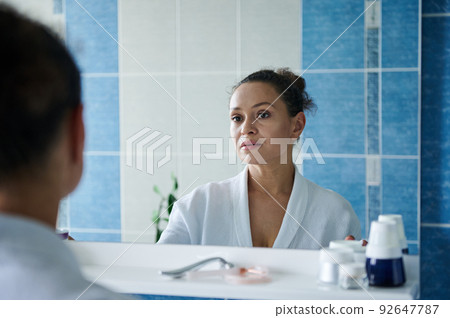 Middle-aged gorgeous woman in bathrobe, looking at her reflection in the bathroom mirror during morning beauty procedure 92647787
