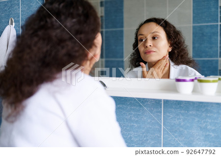 Charming 35s young woman in a white bathrobe examines her face in the mirror, for the first signs of aging and wrinkles. 92647792