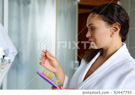 Middle aged brunette, in a white bathrobe inspecting the lost hair on a hairbrush. Problems of hair loss and splits end 92647795