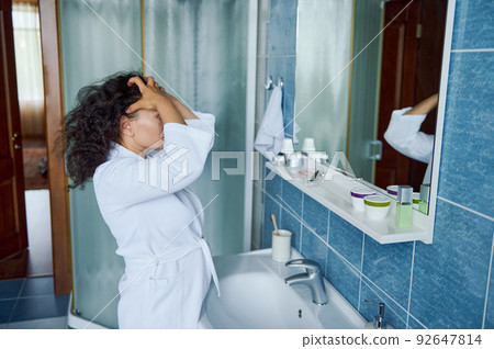 Cute woman in white bathrobe standing in bathroom reflected in mirror, touching curly hair during morning beauty routine Cute woman in white bathrobe standing in bathroom reflected in mirror, touching curly hair during morning beauty routine 92647814