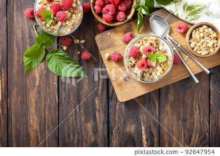 Healthy breakfast menu concept. Home made granola breakfast. Glass of parfait made of granola, berries raspberry, yogurt with chia seeds on rustic table. View from above. Copy space. Healthy breakfast menu concept. Home made granola breakfast. Glass of parfait made of granola, berries raspberry, yogurt with chia seeds on rustic table. View from above. Copy space. 92647954
