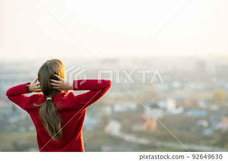 Young woman in red jacket standing with her hands behind head outdoors enjoying evening view. Relaxing, freedom and wellness concept. Young woman in red jacket standing with her hands behind head outdoors enjoying evening view. Relaxing, freedom and wellness concept. 92649630