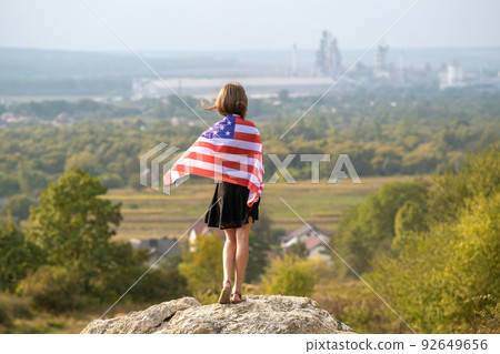 Young pretty american woman with long hair holding waving on wind USA flag on her sholders standing outdoors enjoying warm summer day. 92649656