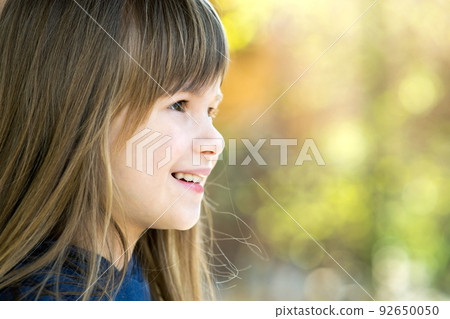 Portrait of pretty child girl with gray eyes and long fair hair smiling outdoors on blurred bright background. Cute female kid on warm summer day outside. 92650050