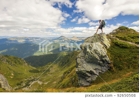 Hiker with a backpack standing o big stone in green rocky mounta Hiker with a backpack standing o big stone in green rocky mounta 92650508