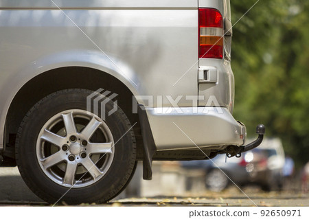 Close-up side view detail of silver passenger medium size luxury minibus van with tow-bar parked on summer sunny city street pavement with blurred silhouettes of pedestrians and cars in background. 92650971