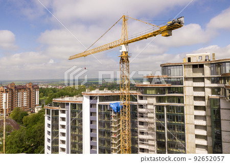 Apartment or office tall building under construction. Brick walls, glass windows, scaffolding and concrete support pillars. Tower crane on bright blue sky copy space background. Apartment or office tall building under construction. Brick walls, glass windows, scaffolding and concrete support pillars. Tower crane on bright blue sky copy space background. 92652057