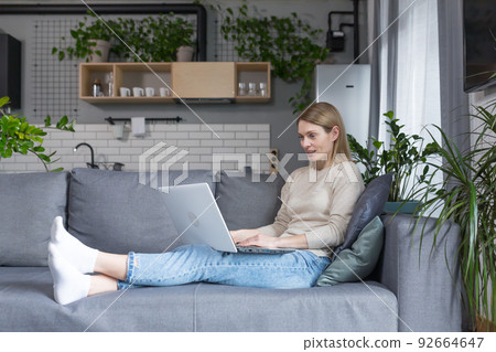 Beautiful woman at home sitting on sofa smiling and working on laptop Beautiful woman at home sitting on sofa smiling and working on laptop 92664647