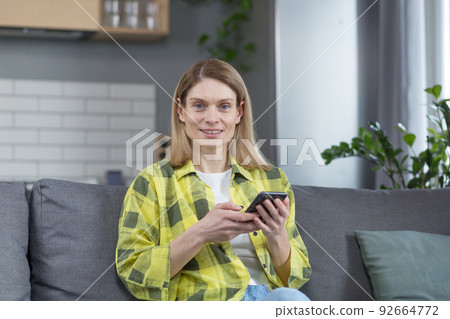 Happy middle-aged blonde woman sitting on sofa at home looking at camera and smiling holding phone in hands Happy middle-aged blonde woman sitting on sofa at home looking at camera and smiling holding phone in hands 92664772