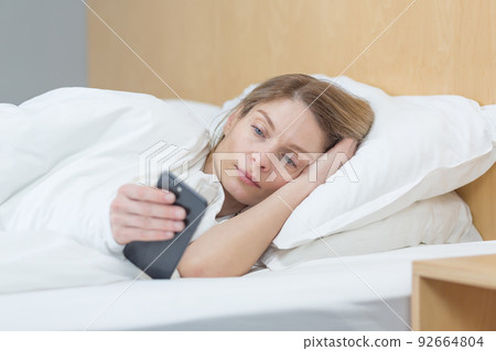 A woman lies under a blanket at home in the morning, looks at the phone screen, reads the morning news 92664804