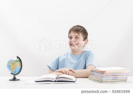 Cheerful laughing teenage boy sits at table while doing his homework. Stack of notebooks and globe on table. International education. Cheerful laughing teenage boy sits at table while doing his homework. Stack of notebooks and globe on table. International education. 92665099