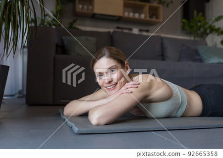 Close-up portrait of fitness woman at home smiling and looking at camera while lying on sports mat 92665358