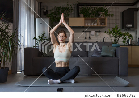 Woman doing yoga at home sitting in lotus position on the floor, using phone for online fitness and yoga classes 92665382