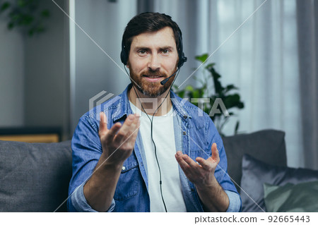 Close-up photo portrait of a bearded man at home with a headset, looking at the camera and talking fun on a video call, looking at the camera sitting at home on the couch Close-up photo portrait of a bearded man at home with a headset, looking at the camera and talking fun on a video call, looking at the camera sitting at home on the couch 92665443