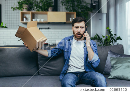 Annoyed and angry man at home received the wrong parcel from the online store, calls in support while sitting on the couch Annoyed and angry man at home received the wrong parcel from the online store, calls in support while sitting on the couch 92665520