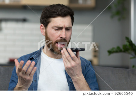 Angry man records audio message, uses app, and dictaphone on phone, sitting on sofa at home 92665540