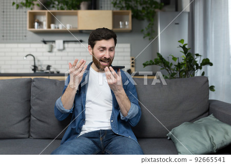 Angry man records audio message, uses app, and dictaphone on phone, sitting on sofa at home 92665541