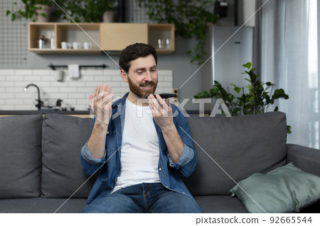 Man at home happily records audio messages, dictates information to the dictaphone of the phone, sitting on the couch at home 92665544
