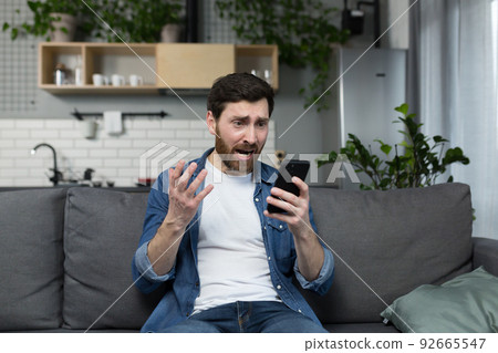The man himself at home, shocked and disappointed, looks at the phone, sitting on the couch The man himself at home, shocked and disappointed, looks at the phone, sitting on the couch 92665547