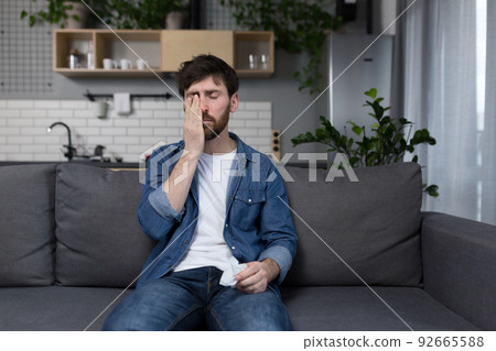 Sick man with a beard sits at home on the couch, has the flu and cold, allergy sneezes with a napkin 92665588