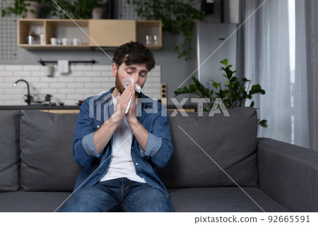The man is sick at home, has the flu and runny nose, sneezes, sits on the couch alone in the kitchen The man is sick at home, has the flu and runny nose, sneezes, sits on the couch alone in the kitchen 92665591