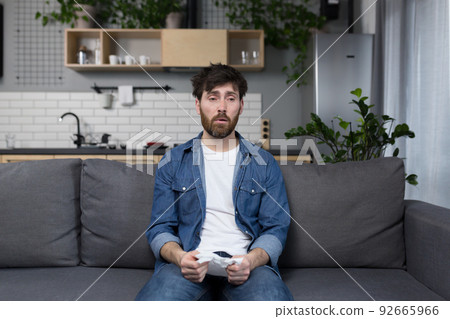 Sick man with a beard sits at home on the couch, has the flu and cold, allergy sneezes with a napkin Sick man with a beard sits at home on the couch, has the flu and cold, allergy sneezes with a napkin 92665966