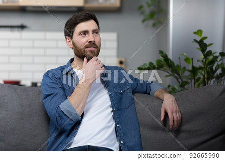 The man at home is sitting on the couch, resting looking away, smiling dreamy pensive The man at home is sitting on the couch, resting looking away, smiling dreamy pensive 92665990