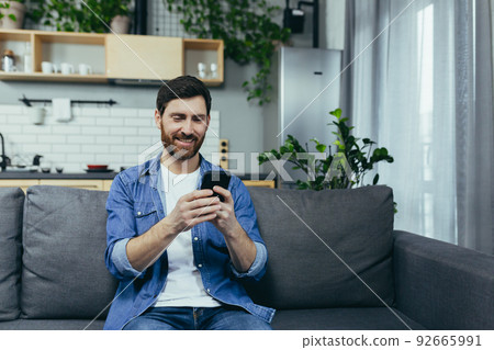 Happy man smiling and using phone sitting on sofa at home Happy man smiling and using phone sitting on sofa at home 92665991