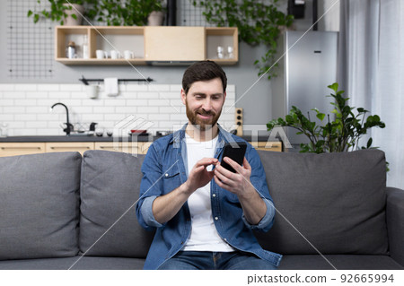 Happy man smiling and using phone sitting on sofa at home Happy man smiling and using phone sitting on sofa at home 92665994